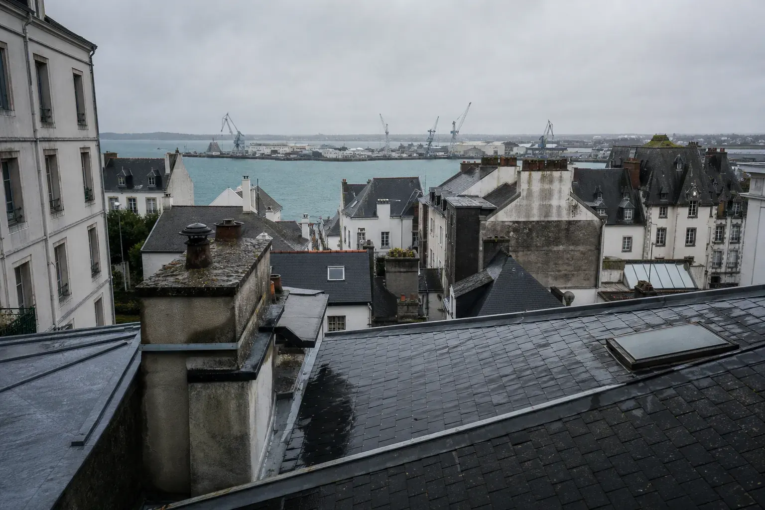 Toits d’ardoise et de tuile mécanique au premier plan, rade et grues portuaires au fond sous un ciel bas